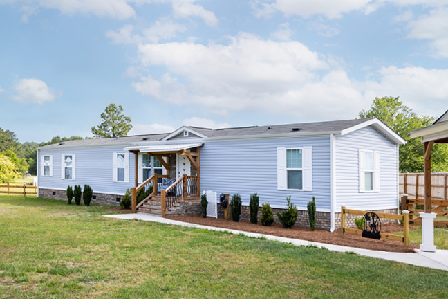 A mobile home with light blue siding and small planted shrubs lining the front.