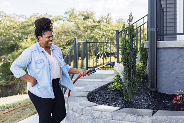 A smiling woman waters the plants outside of her manufactured home.