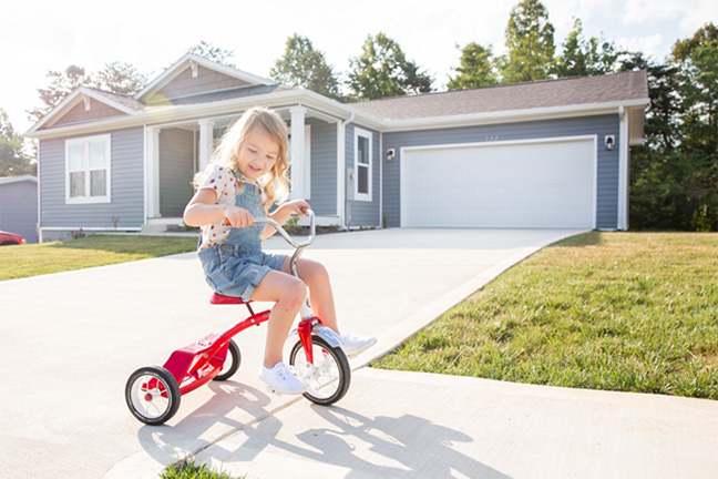 A girl on a tricycle with a blue-exterior manufactured home at the top of the driveway.
