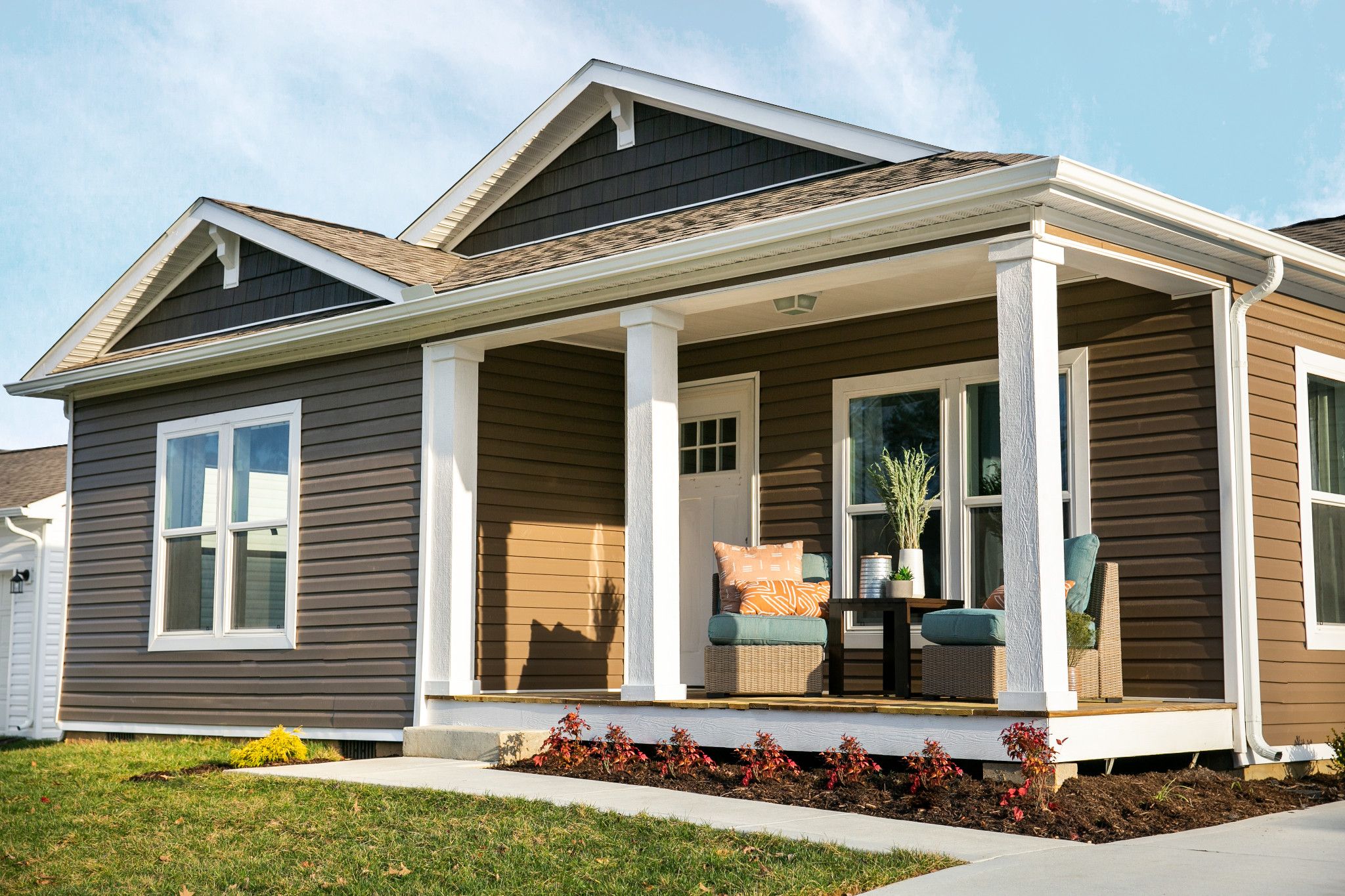 A modular home with brown vinyl siding, white columns, front porch furniture and a small lawn. Modular housing can be indistinguishable from stick-built homes, depending on its design.