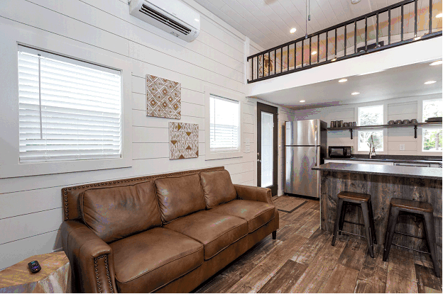  The interior of a manufactured home in a farmhouse style. The floors are covered with thick wooden floor planks that have a rustic look. The walls are covered in shiplap painted white to make the interior of the mobile home look brighter.