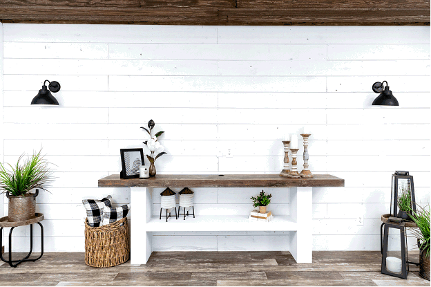 The interior of a mobile home in the style of a farmhouse. The walls are covered in shiplap and painted white to help light reflect throughout the home. A rustic sideboard with a live-edge wooden table top stands against the wall. A natural wooden beam runs across the top of the wall.