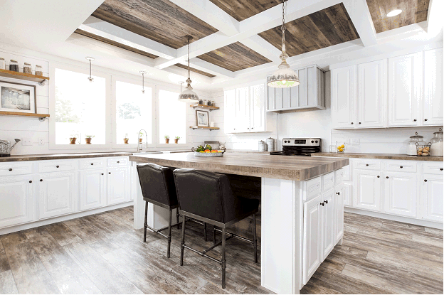 The interior of a rustic farmhouse style kitchen in a manufactured home. The kitchen island and counters feature natural wooden countertops that match the rustic wood flooring. Natural wooden shiplap is also featured on the ceiling, along with thick white accent beams. Three large, bright windows allow plenty of light into the kitchen of the mobile home. 