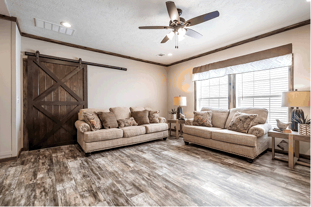 A large, natural wooden barn door stands along the wall. The floors are made of rustic wood planks. A neutral beige sofa and loveseat are featured next to two large windows with rustic wooden window trim.