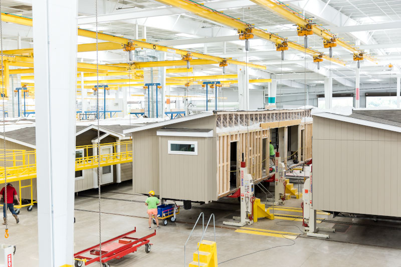 A group of workers builds manufactured homes in a large warehouse. Modular or manufactured homes are created off-site and then shipped to a plot of land.