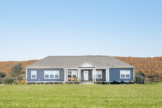 The exterior of a brand-new modular home with a large front yard. The house has blue vinyl siding with a stone facade. A clear blue sky is overhead, and mountains rise in the distance.