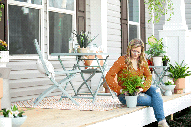 A young woman sits on the front porch of her home as she tends to her plants. The porch features many potted plants, an outdoor rug, and a set of blue metal chairs and matching table. A bottle of white wine with two glasses sits on top of the table.