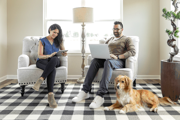 A young couple browse their electronic devices while relaxing in their new home. The man is looking on his laptop while the woman browses on a tablet. A dog sits at the man’s feet.
